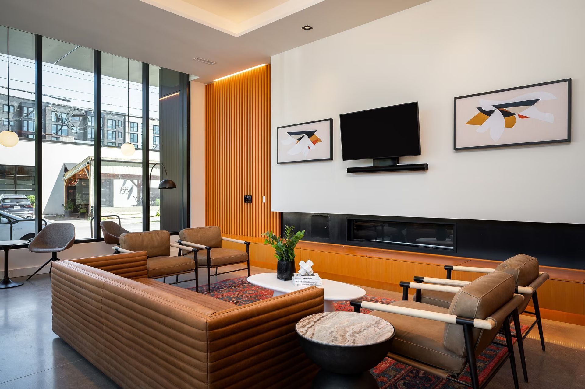Modern living room with a brown sofa, chairs, fireplace, TV, and artwork. Large windows.