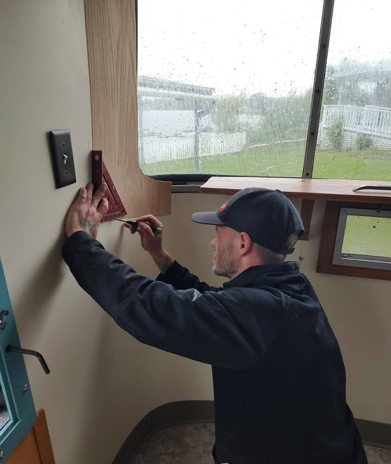Man in a cap wiring a wall-mounted device inside a room beside a window.