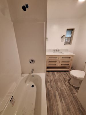 Narrow bathroom with bathtub, wood vanity, toilet, and window on the right.