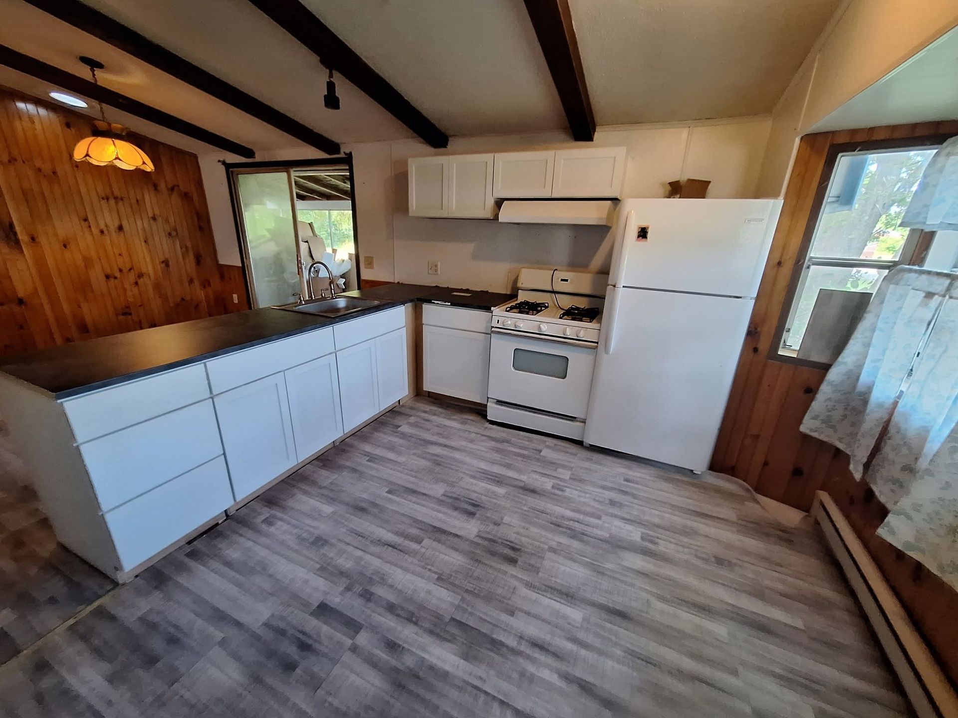 Small rustic kitchen with white cabinets, wood-paneled walls, stove, fridge, and window curtains.
