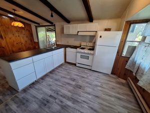 Small rustic kitchen with white cabinets, stove, fridge, wood paneling, and a window with curtains.