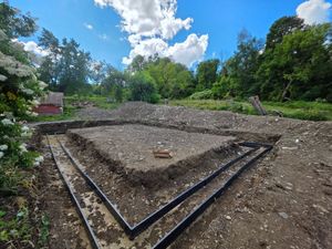 Stone foundation outline in a dirt clearing, surrounded by trees under a blue sky.