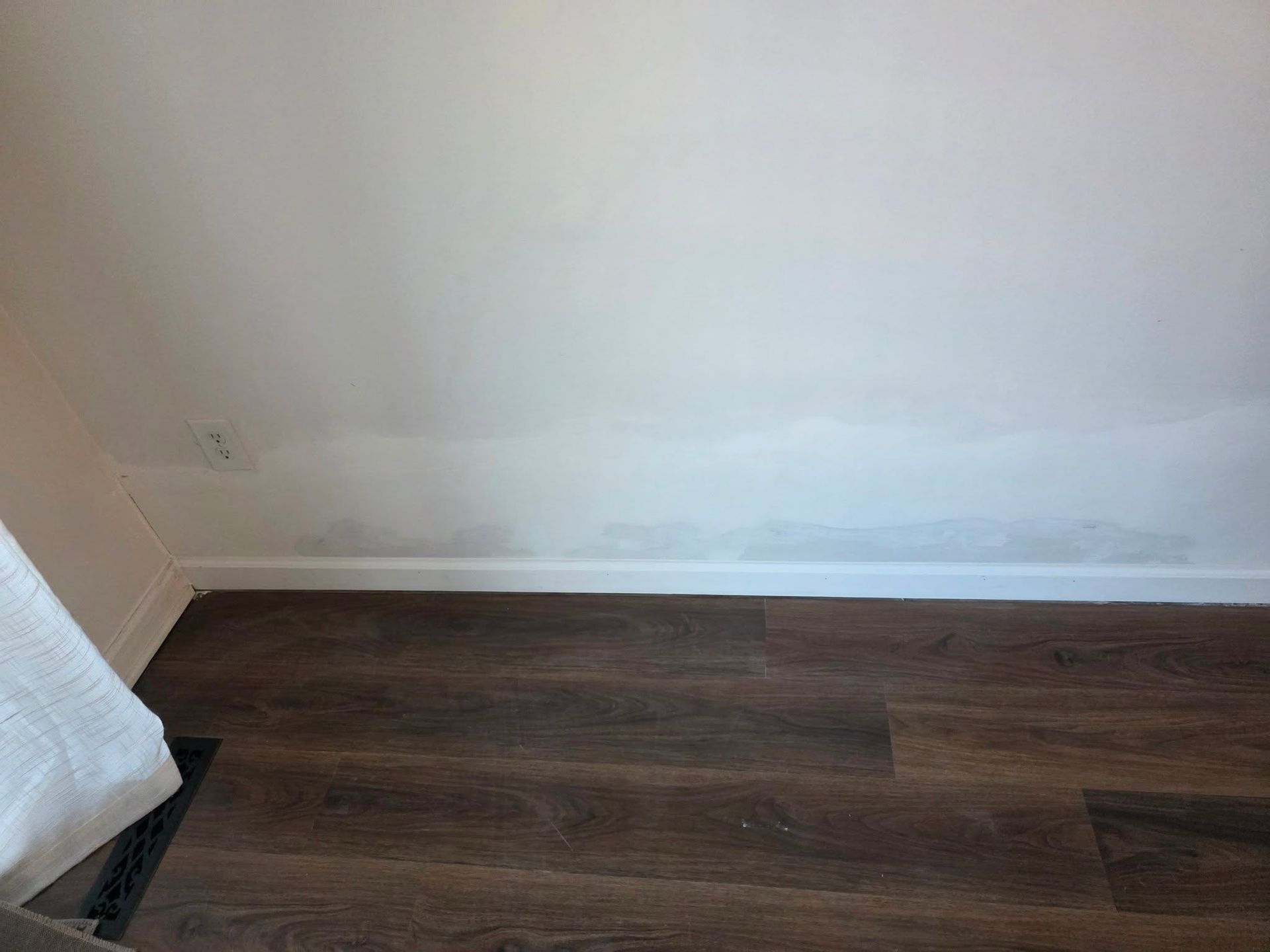 Empty corner of a room with white walls and dark wood flooring, showing a baseboard and window curtain edge