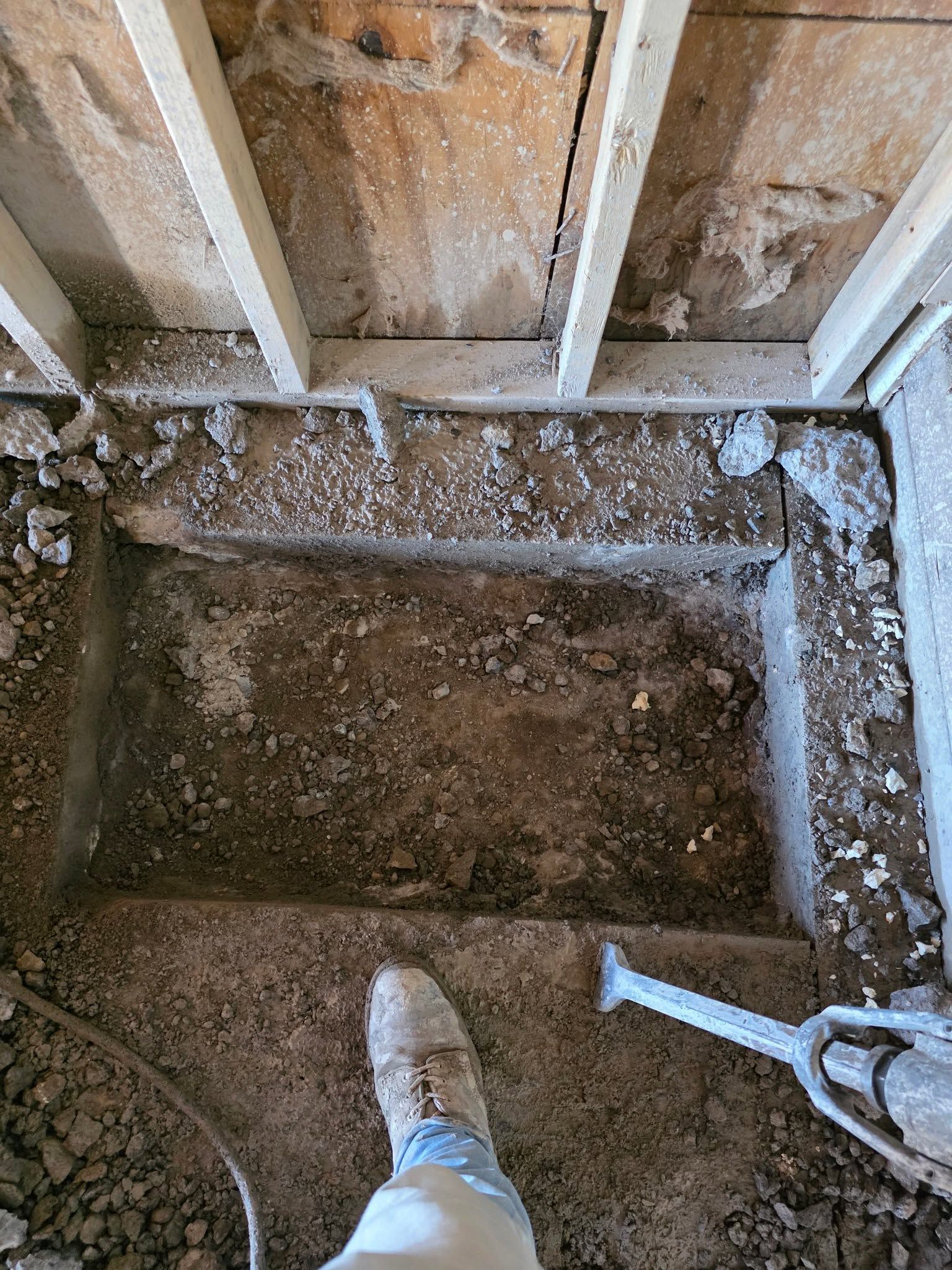 Construction pit in a partially demolished room, with exposed studs, dirt floor, and a shovel visible.