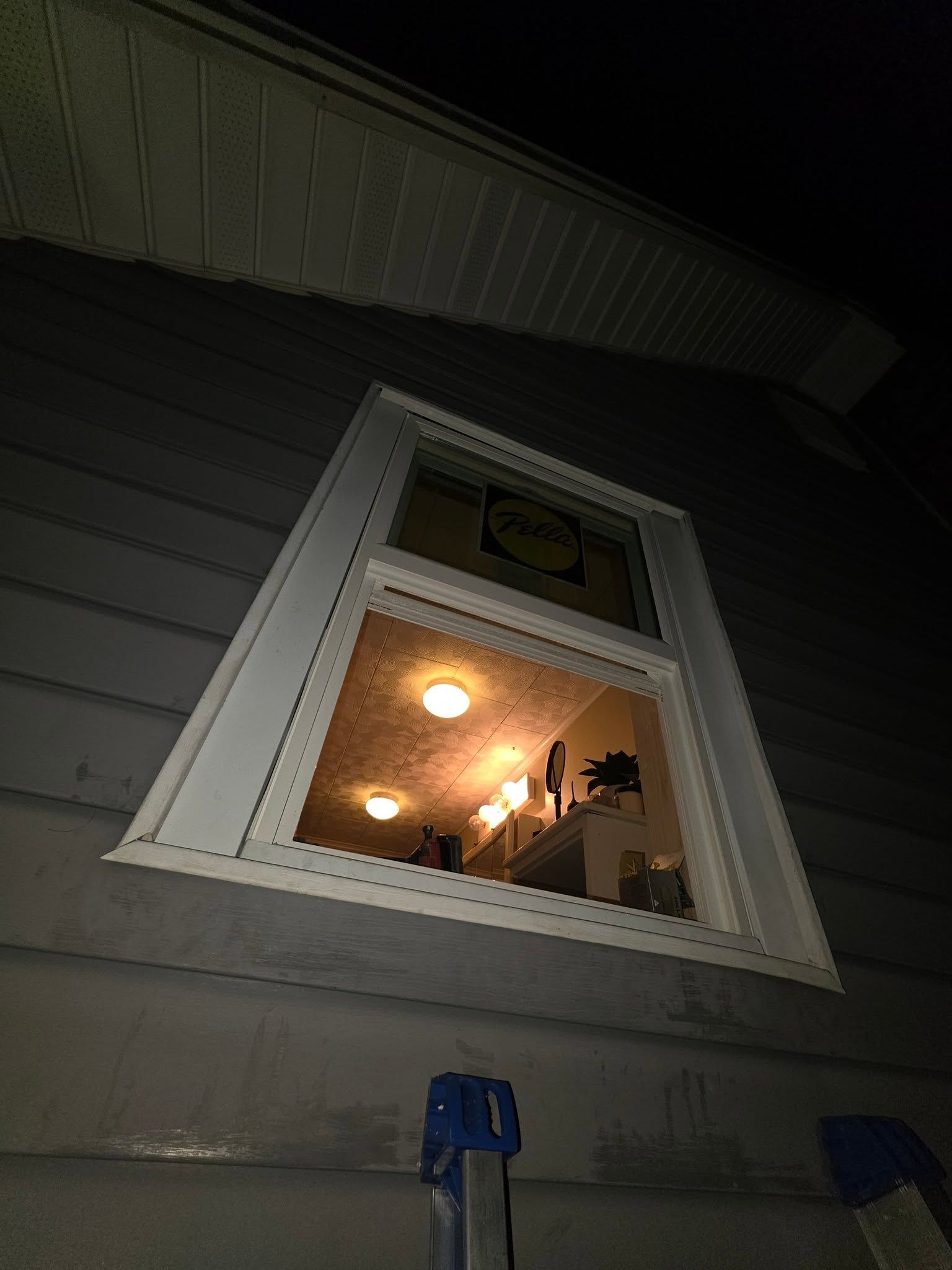 Tilted view of a lit house window at night with white trim and gray siding