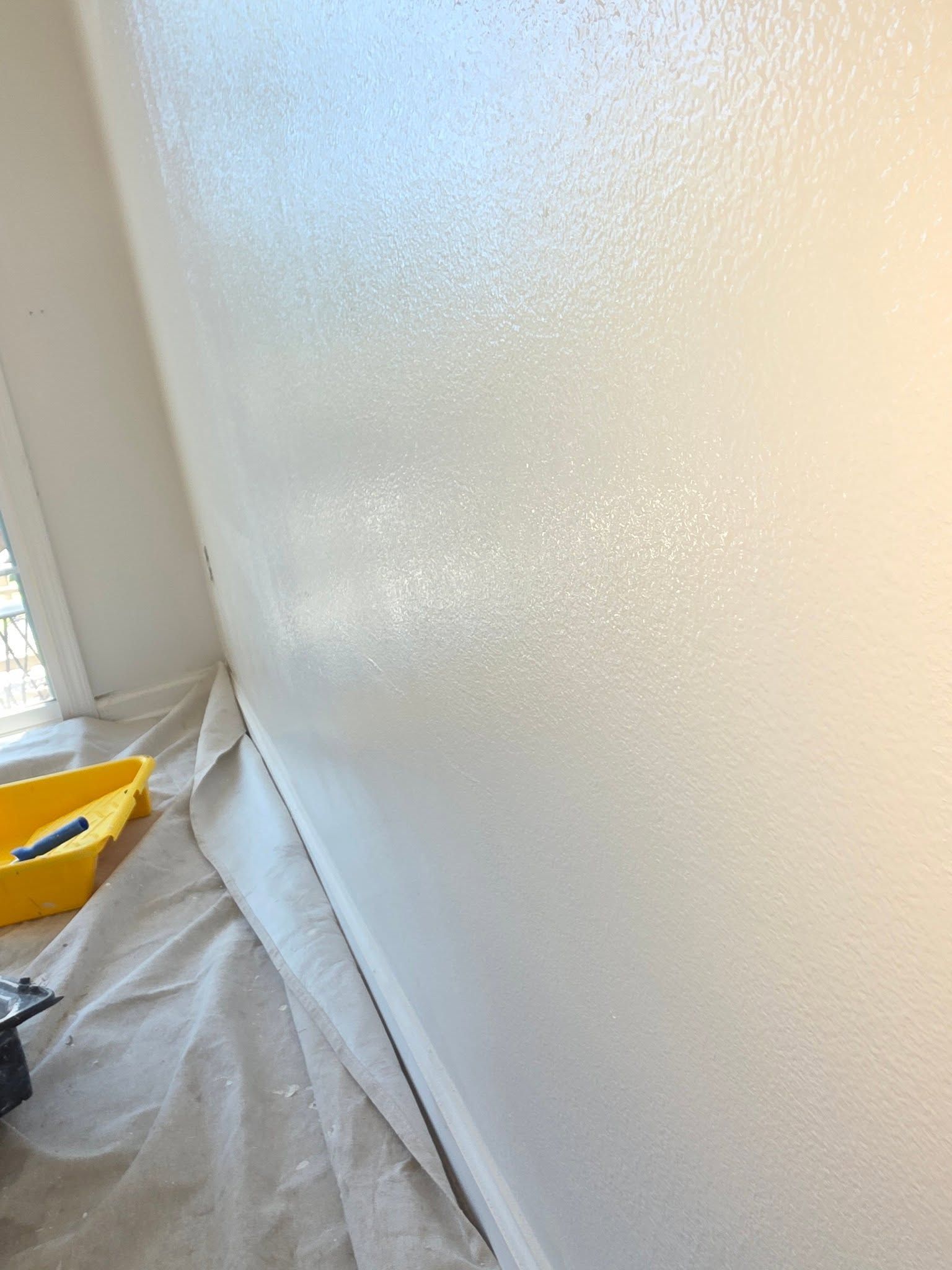 Painted white wall corner beside a window, with a drop cloth and yellow paint tray on the floor.