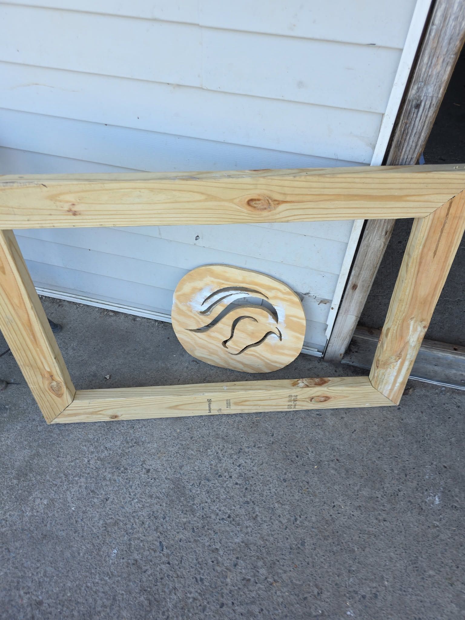 Wooden frame with a circular emblem leaning against a white-sided wall on concrete.