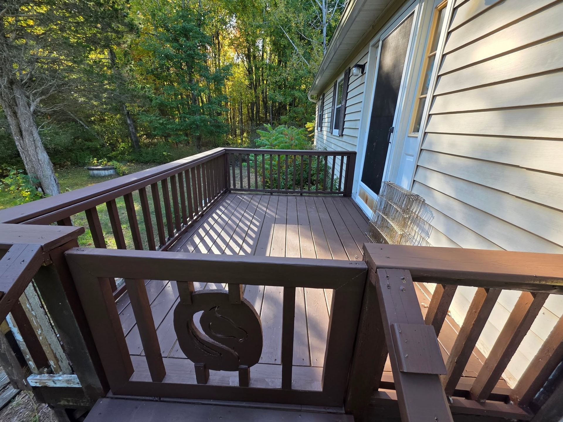 Wooden deck beside a beige house, bordered by dark railing and surrounded by trees.