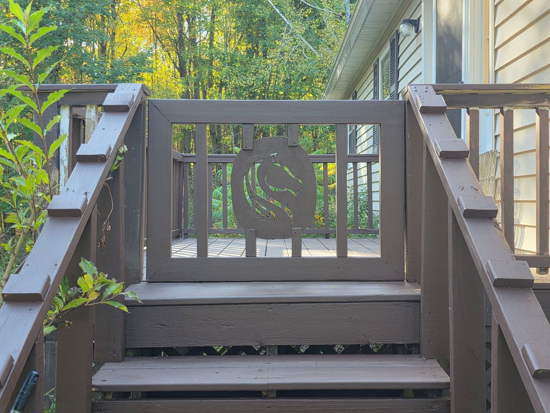 Short outdoor wooden staircase with dark railing and decorative gate, surrounded by greenery