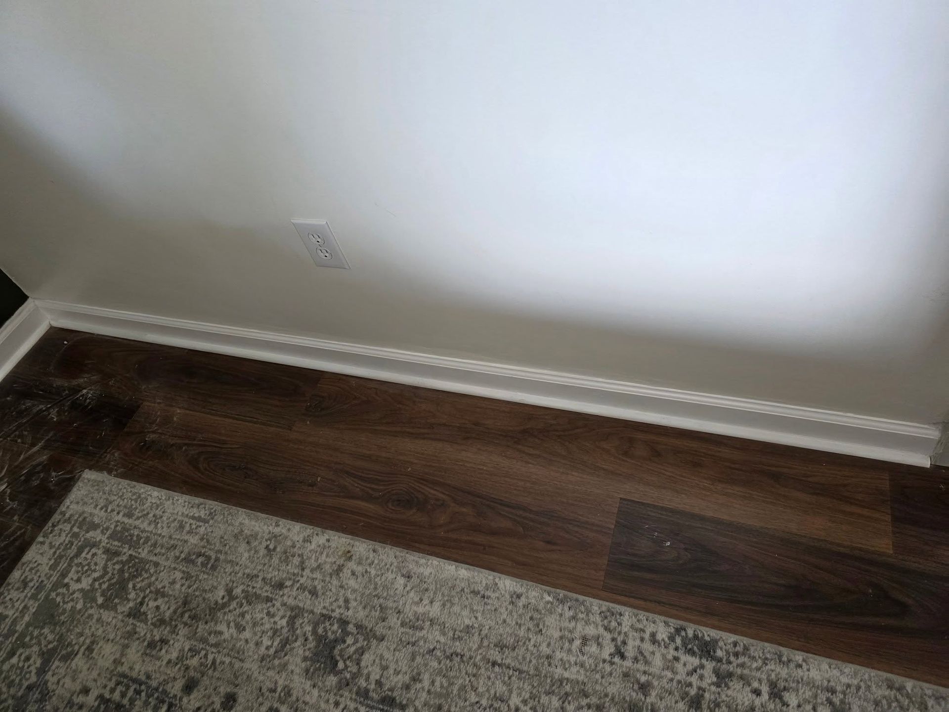 Corner of a room with white wall, hardwood floor, baseboard trim, and part of a gray rug.