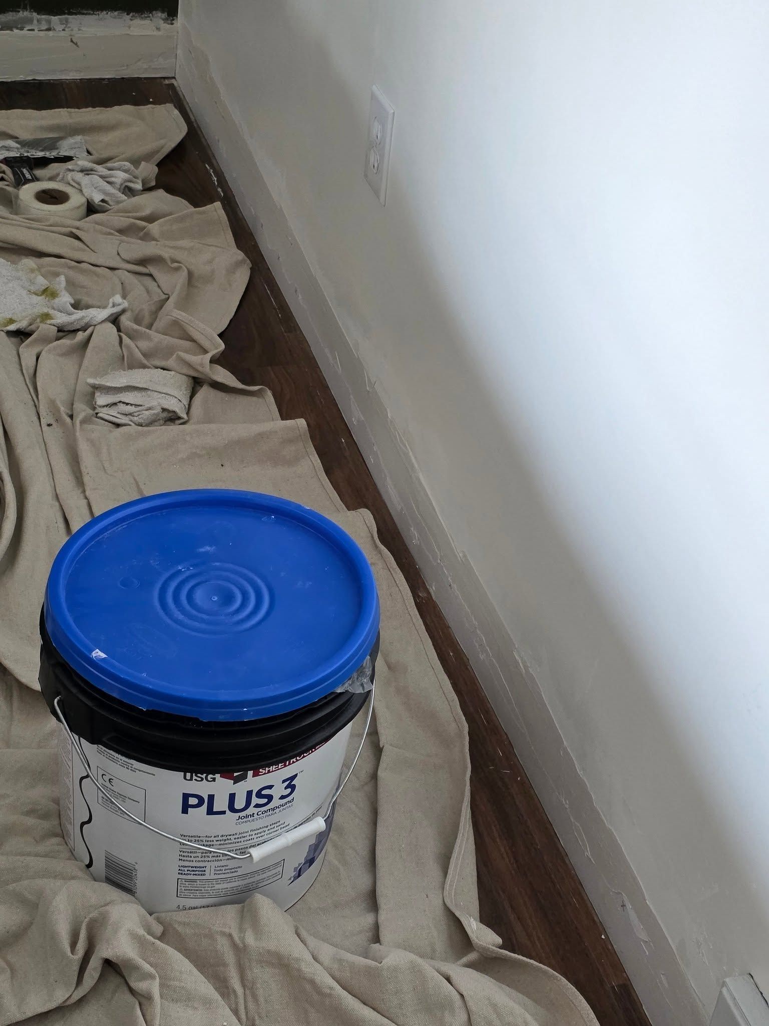 Blue-lidded bucket of paint or sealant on a drop cloth beside a wall in a room under repair