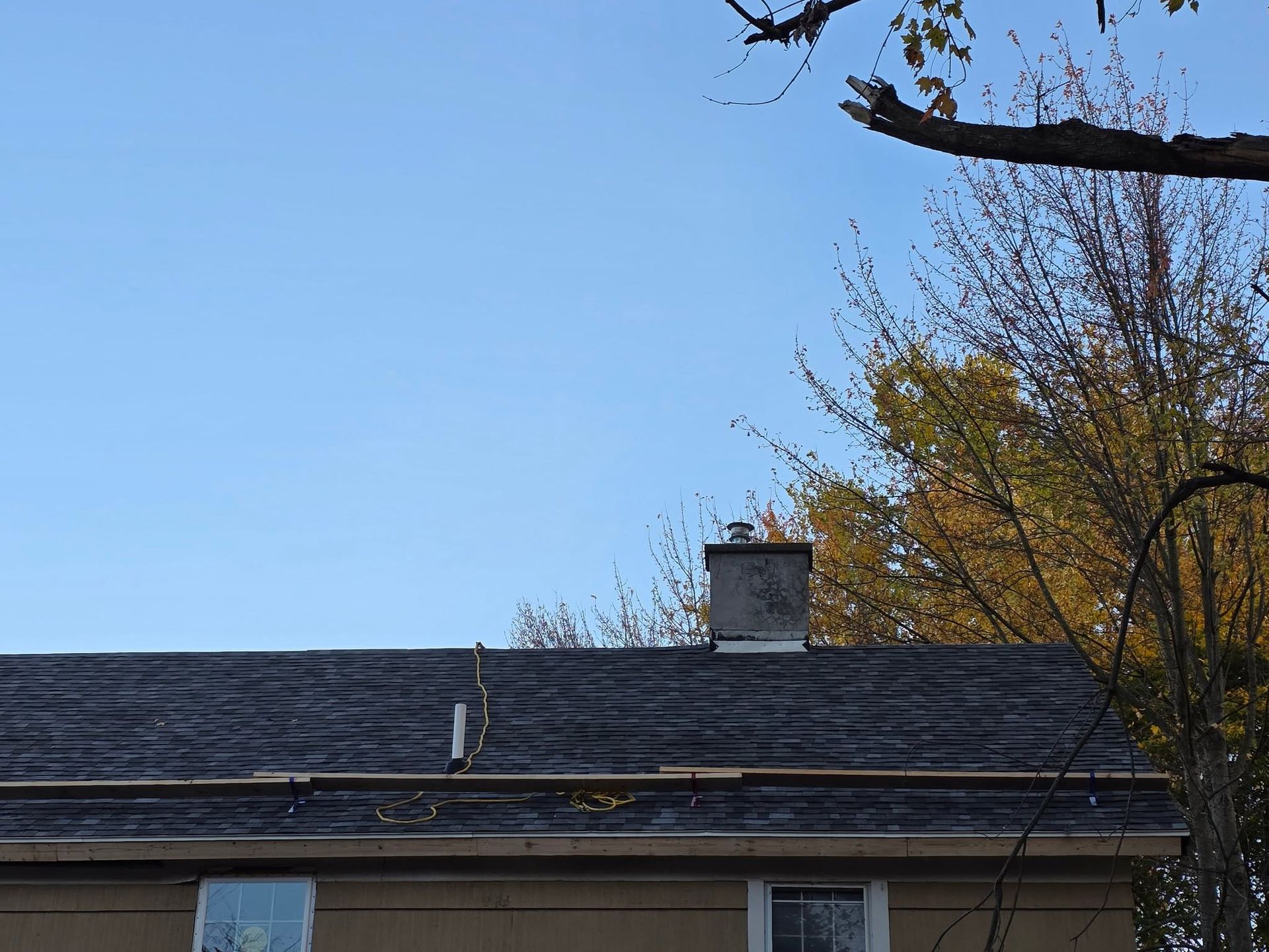 Rooftop with dark shingles and chimney under a clear blue sky, framed by autumn tree branches