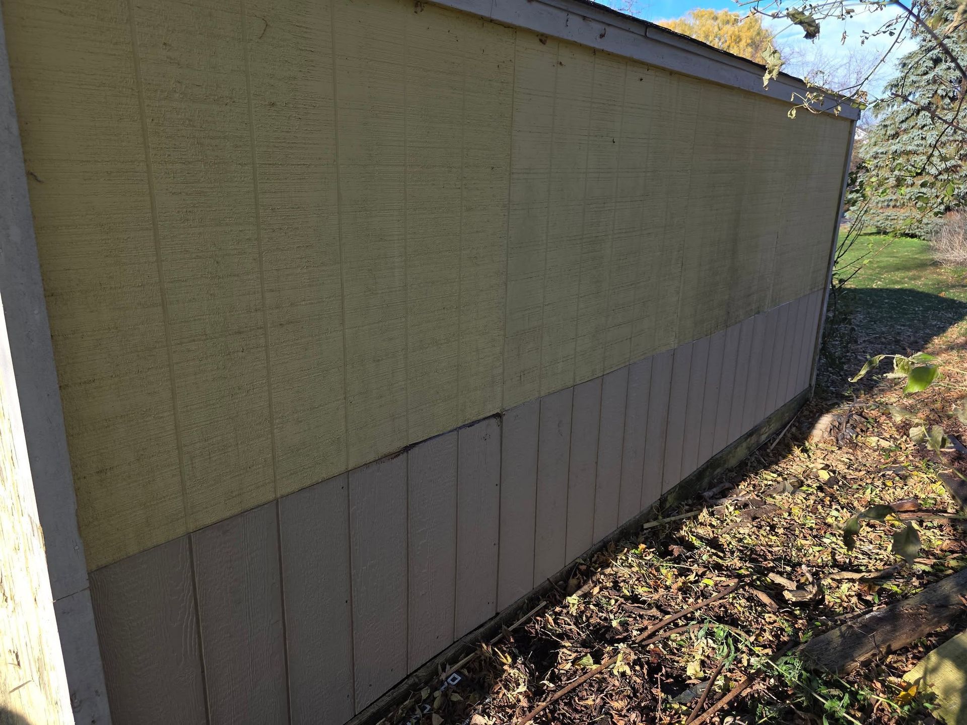 Side of a small building with tan and gray siding beside a grassy, leaf-covered slope.