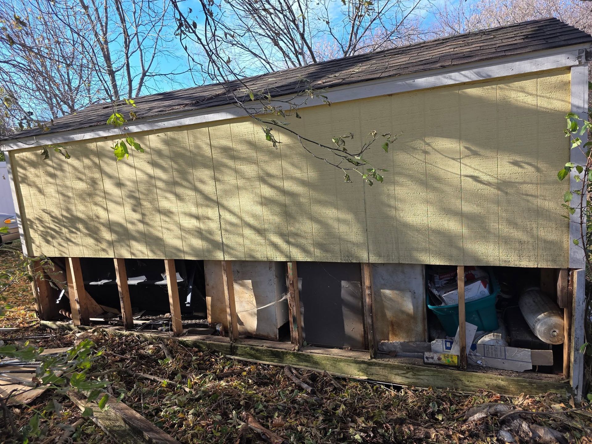Yellow house with lower wall exposed, showing studs and debris in a yard under bare trees