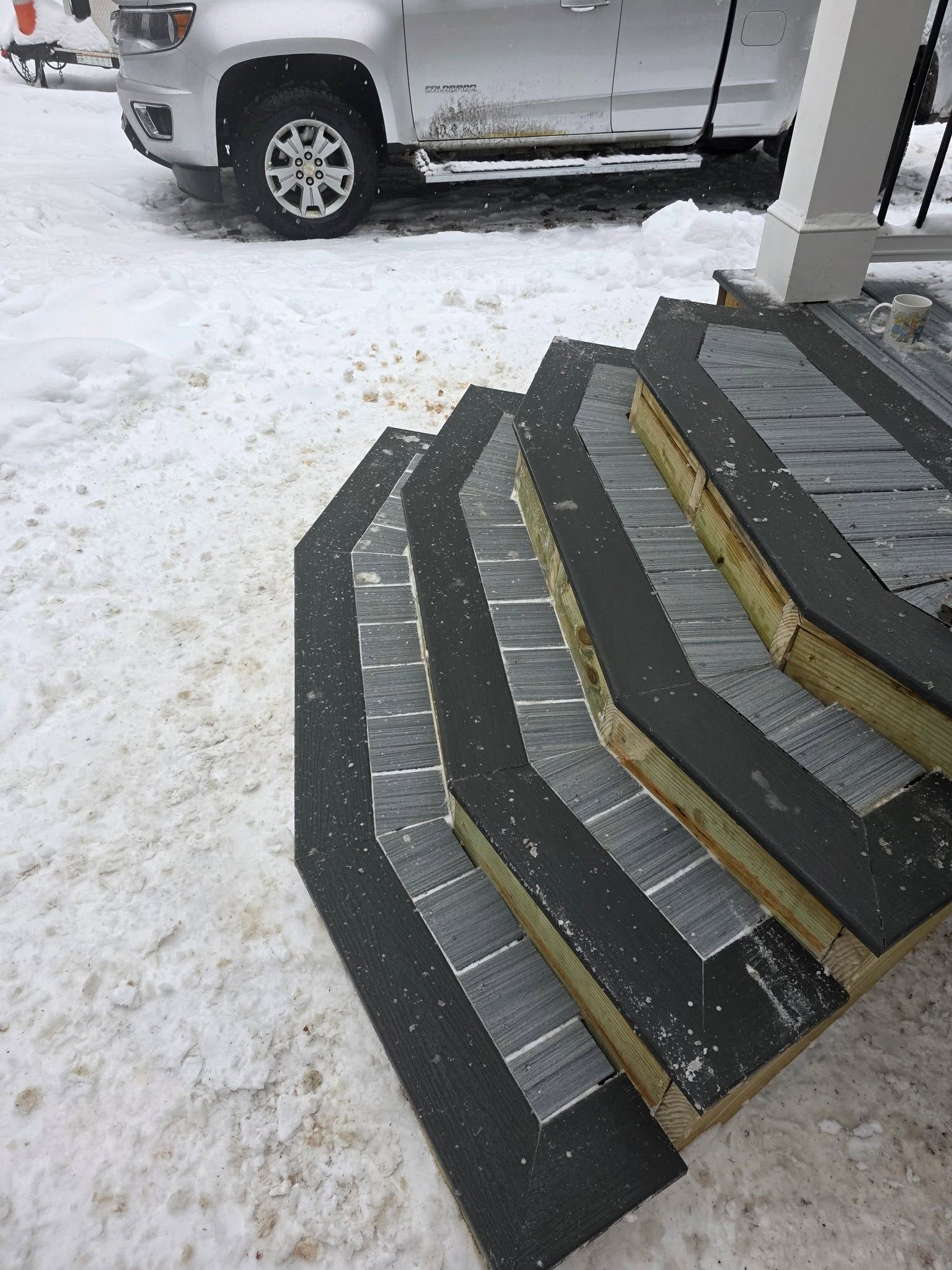 Snow-covered front steps with dark treads and yellow edge strips beside a parked white truck.