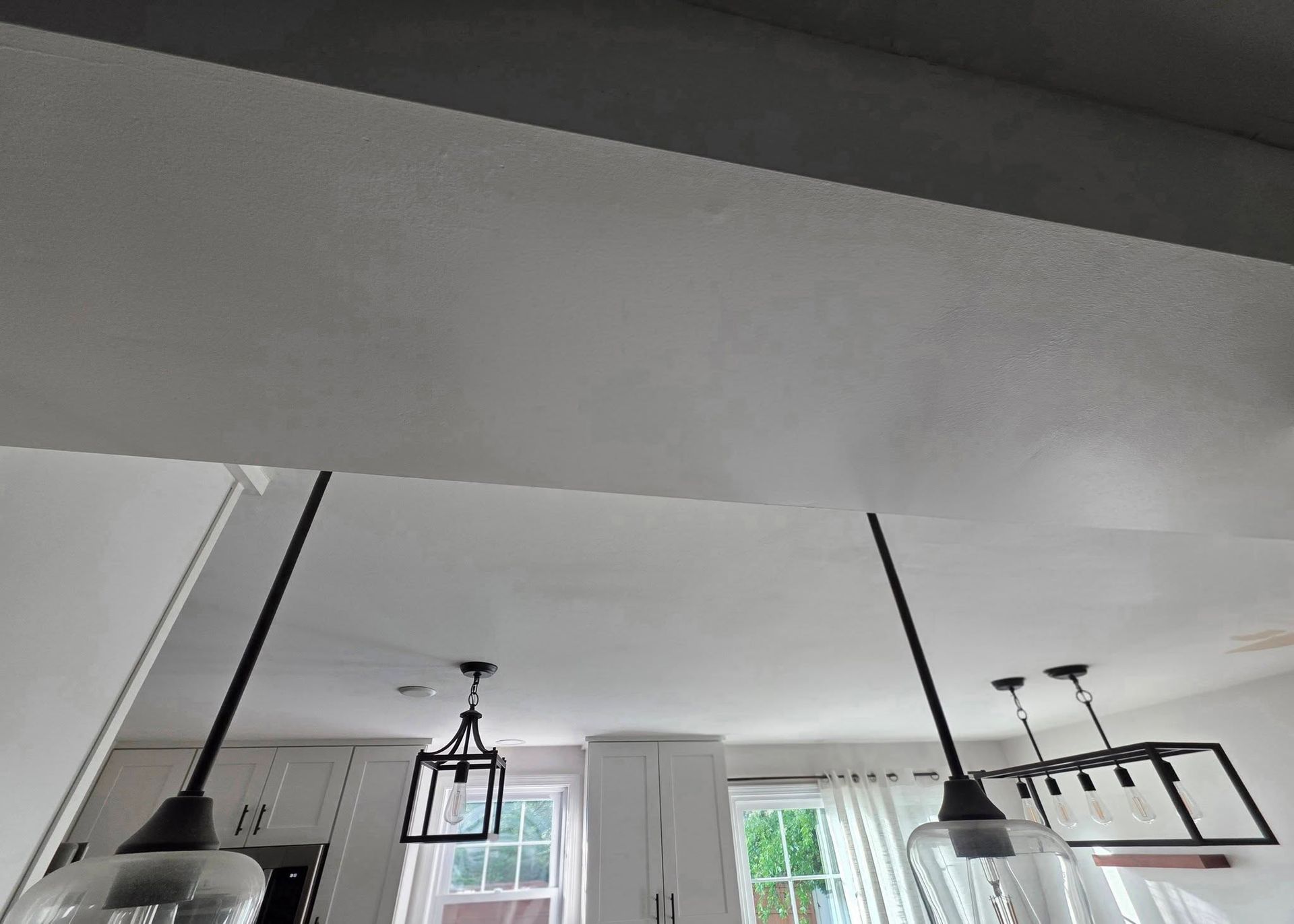 Modern kitchen ceiling with black pendant lights and a rectangular hanging rack above a windowed wall
