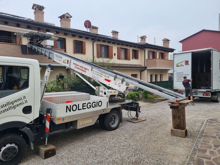Due camion bianchi con piattaforma elevatrice in un cortile ghiaioso vicino a un edificio, uno dei quali contrassegnato con la scritta 