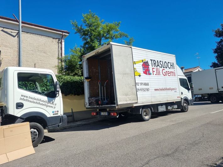 Un camion per traslochi è parcheggiato in una strada soleggiata accanto a un edificio beige, con un altro camion sullo sfondo.