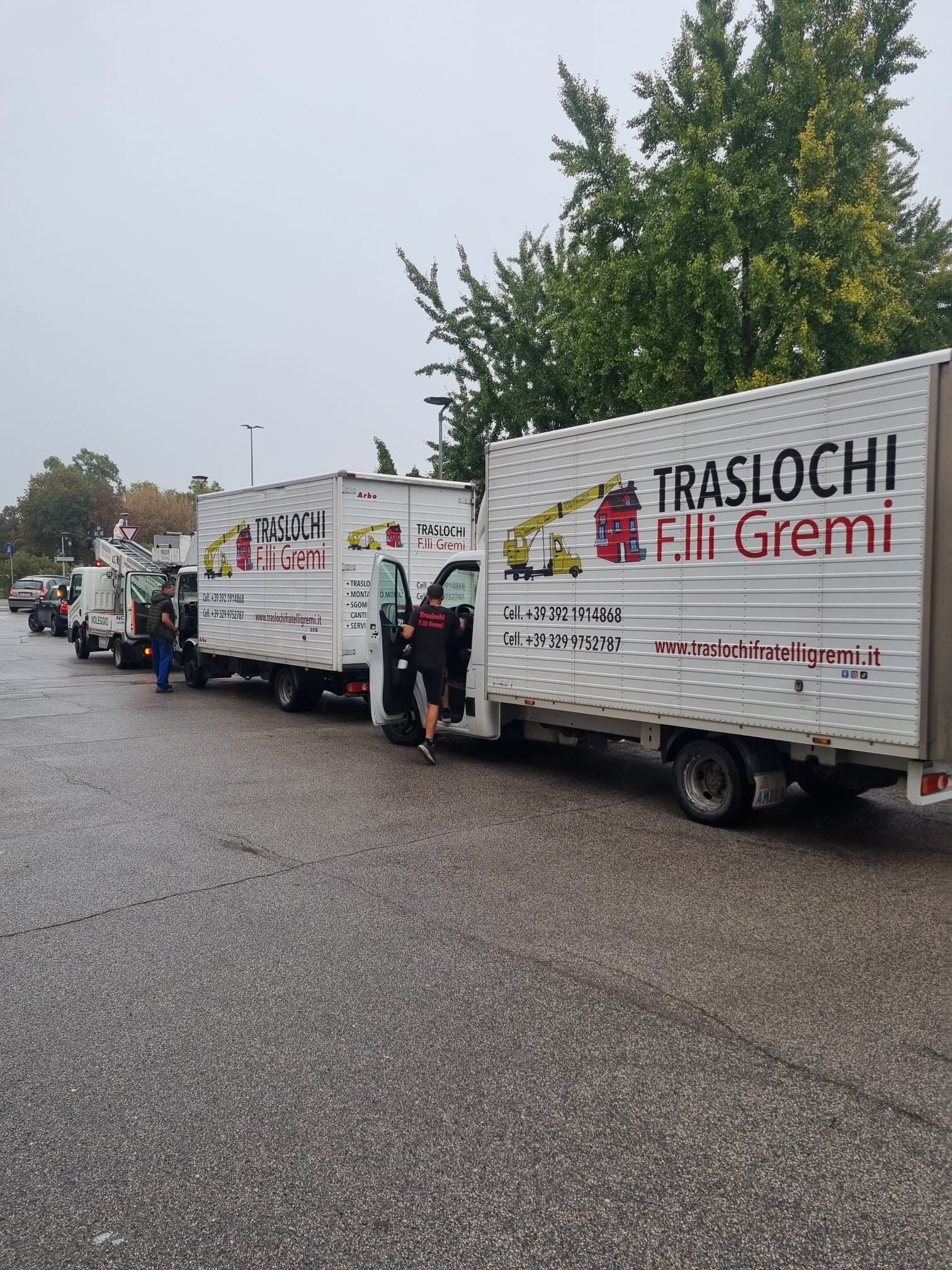 Una fila di camion per traslochi bianchi parcheggiati lungo un ciglio di strada bagnato sotto gli alberi, uno con la scritta