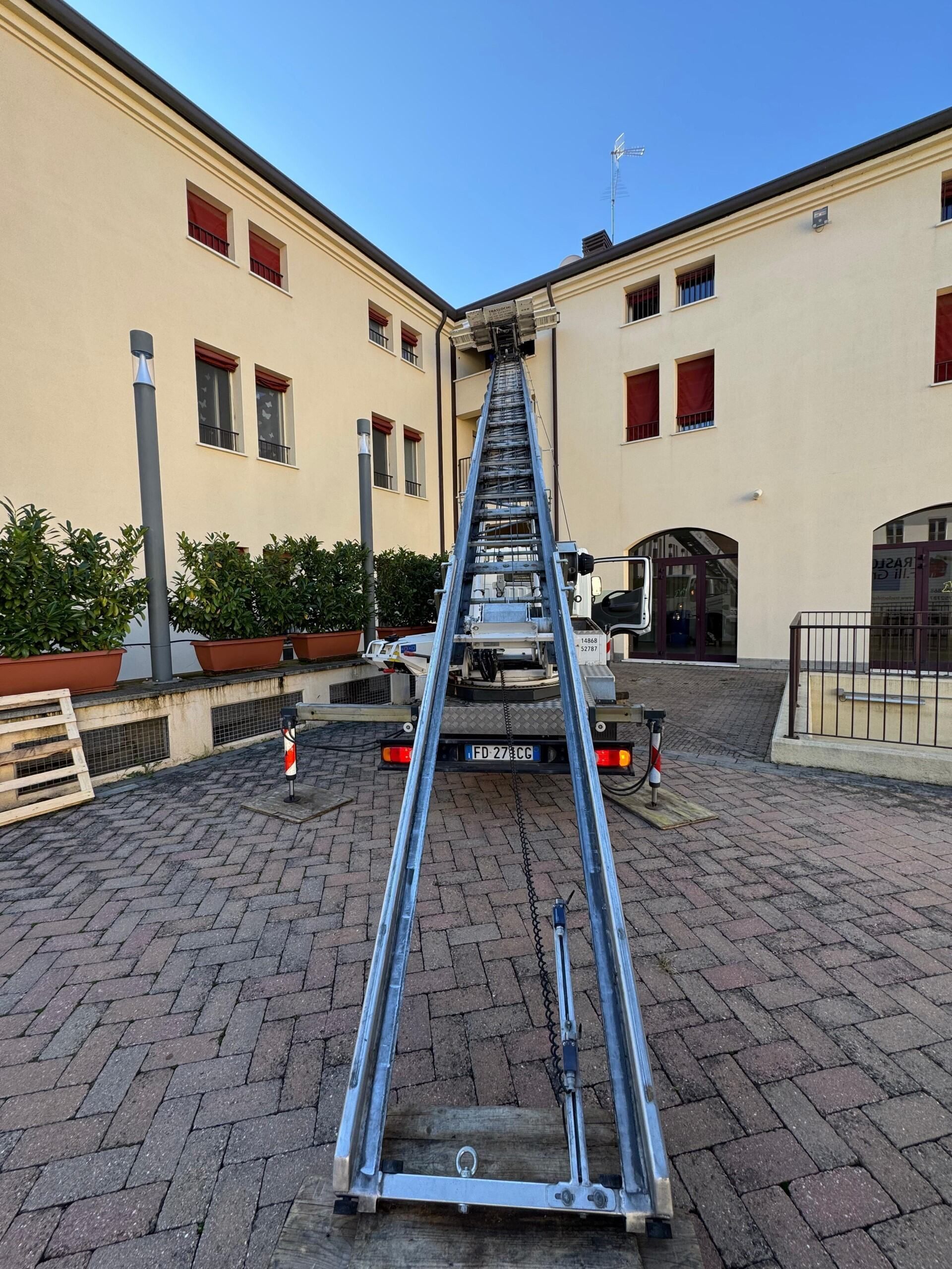 Una scala metallica blu in un cortile, puntata verso l'alto tra due edifici color crema con persiane rosse.