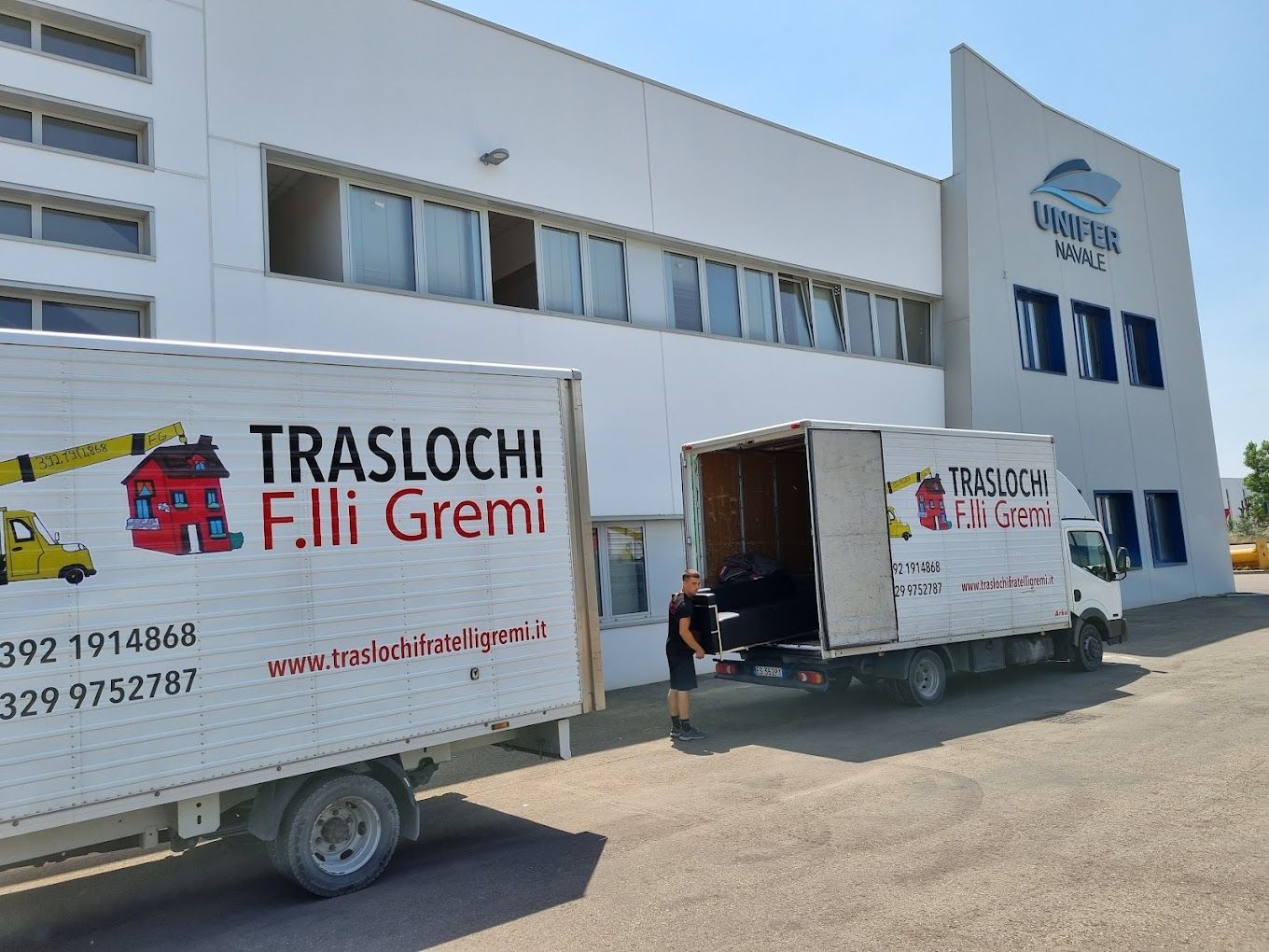 Due camion per traslochi parcheggiati all'esterno di un edificio industriale bianco con una banchina di carico e un logo.