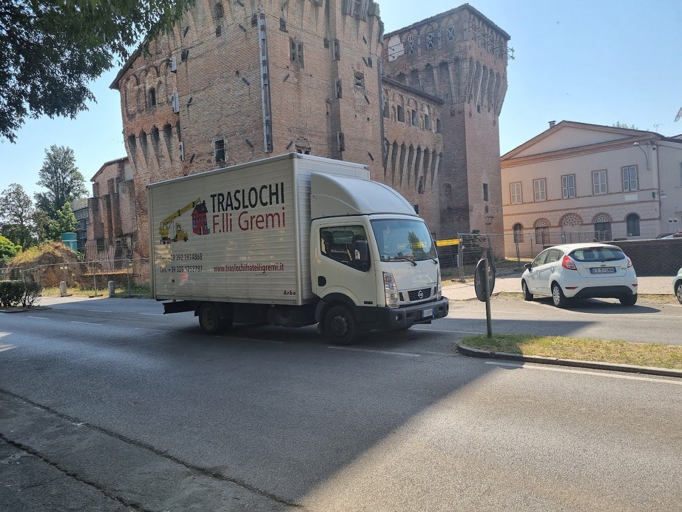 Un camioncino bianco per le consegne svolta in una strada soleggiata vicino a edifici in mattoni e auto parcheggiate.