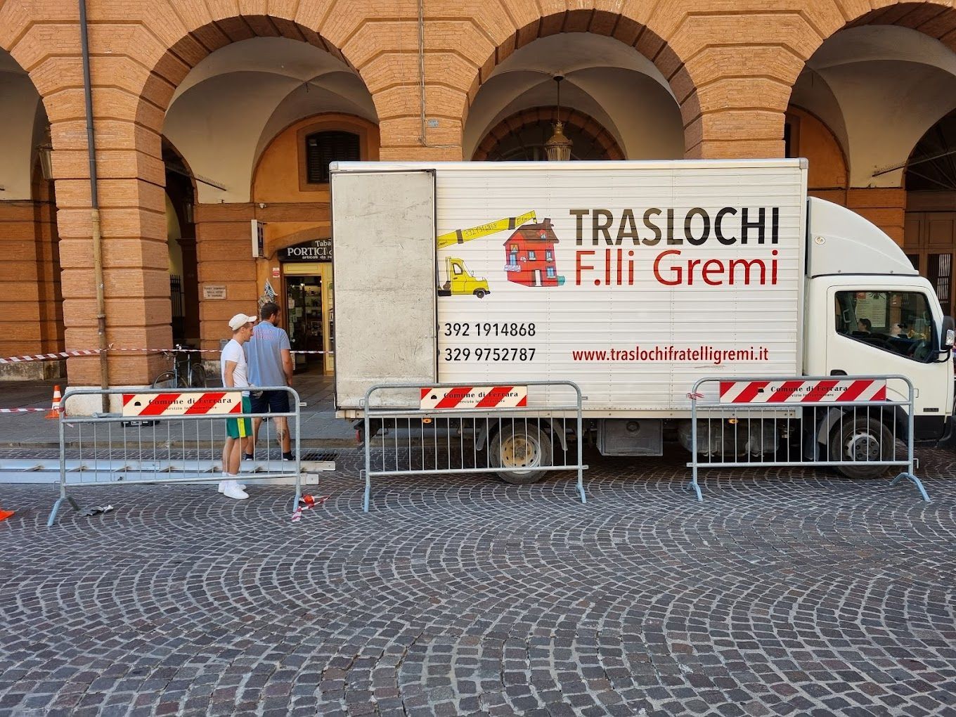 Camion bianco con la scritta “TRASLOCHI Fili Gremi” parcheggiato vicino a un porticato in mattoni, dietro barriere metalliche su una strada acciottolata.
