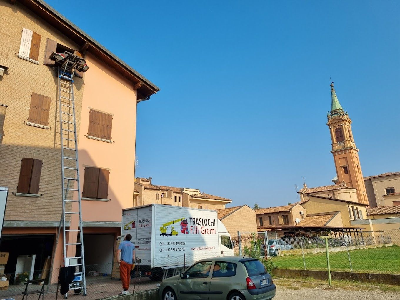 Veduta stradale di un edificio color pesca, auto parcheggiate e un campanile sotto un cielo azzurro limpido.