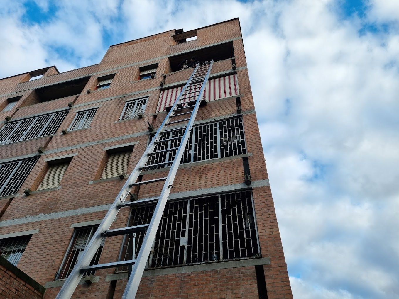 Alto edificio in mattoni con una lunga scala appoggiata a una finestra superiore con le sbarre, stagliata contro un cielo azzurro.