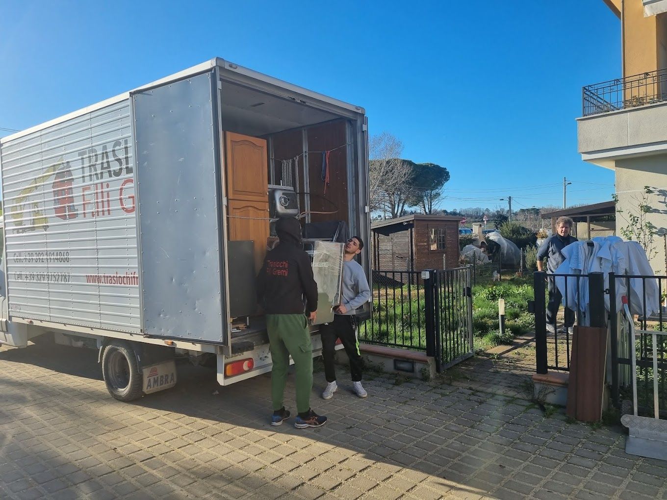 Persone che caricano oggetti su un furgone accanto a una recinzione in una strada soleggiata