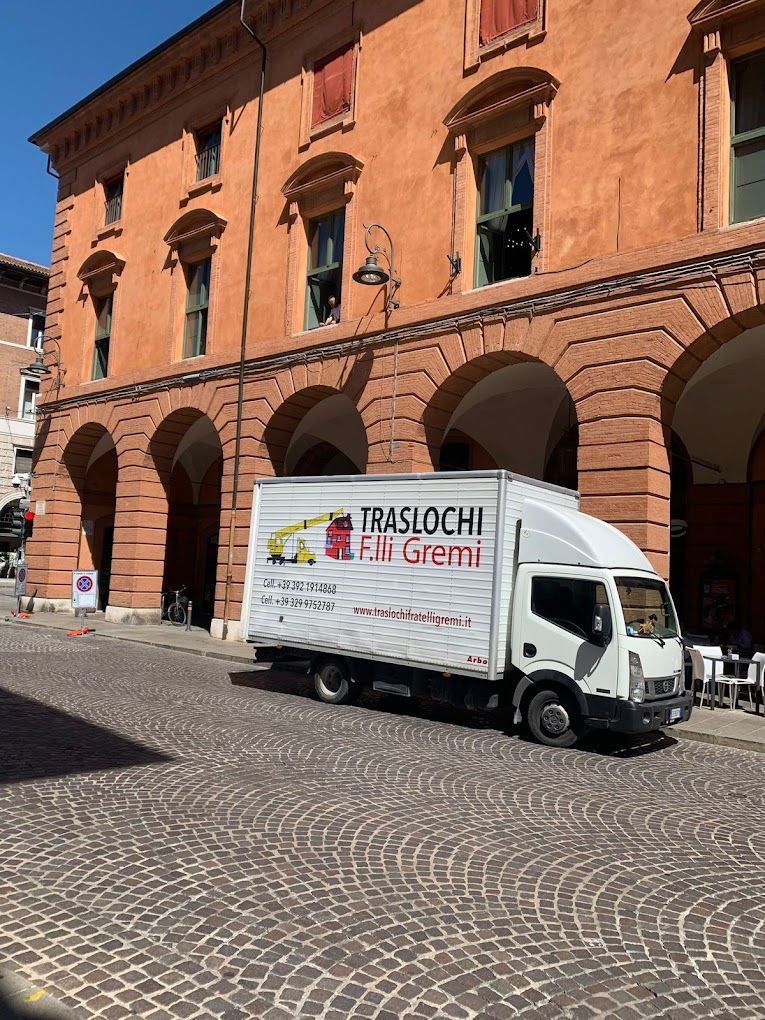 Un furgone bianco per le consegne è parcheggiato accanto a un edificio in mattoni arancioni con archi, su una strada acciottolata.