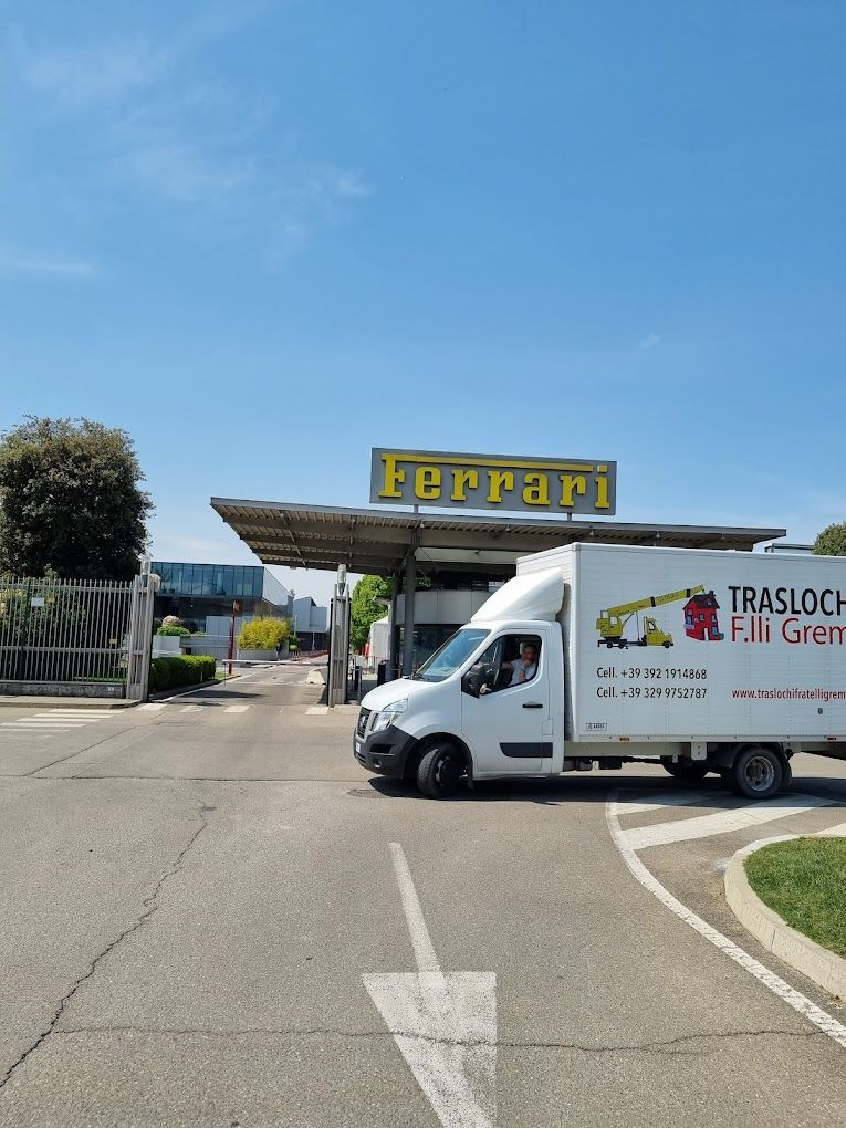 Un furgone bianco per le consegne è parcheggiato davanti all'insegna d'ingresso della Ferrari, su una strada soleggiata.