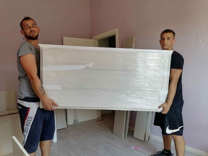 Due uomini trasportano un grande materasso imballato attraverso una stanza con pareti rosa