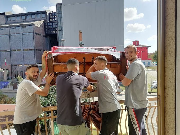 Quattro uomini trasportano un grosso oggetto di legno all'aperto, vicino a un edificio in costruzione.