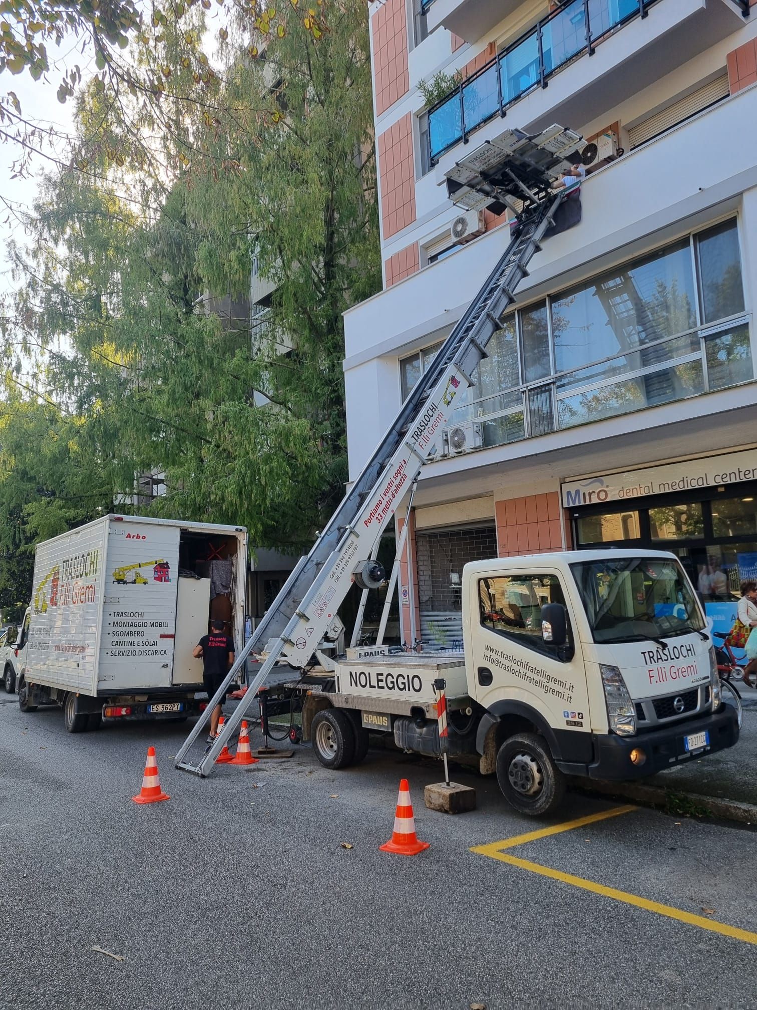Un carro attrezzi utilizza una scala per raggiungere il balcone di un edificio, con coni e un furgone in strada.