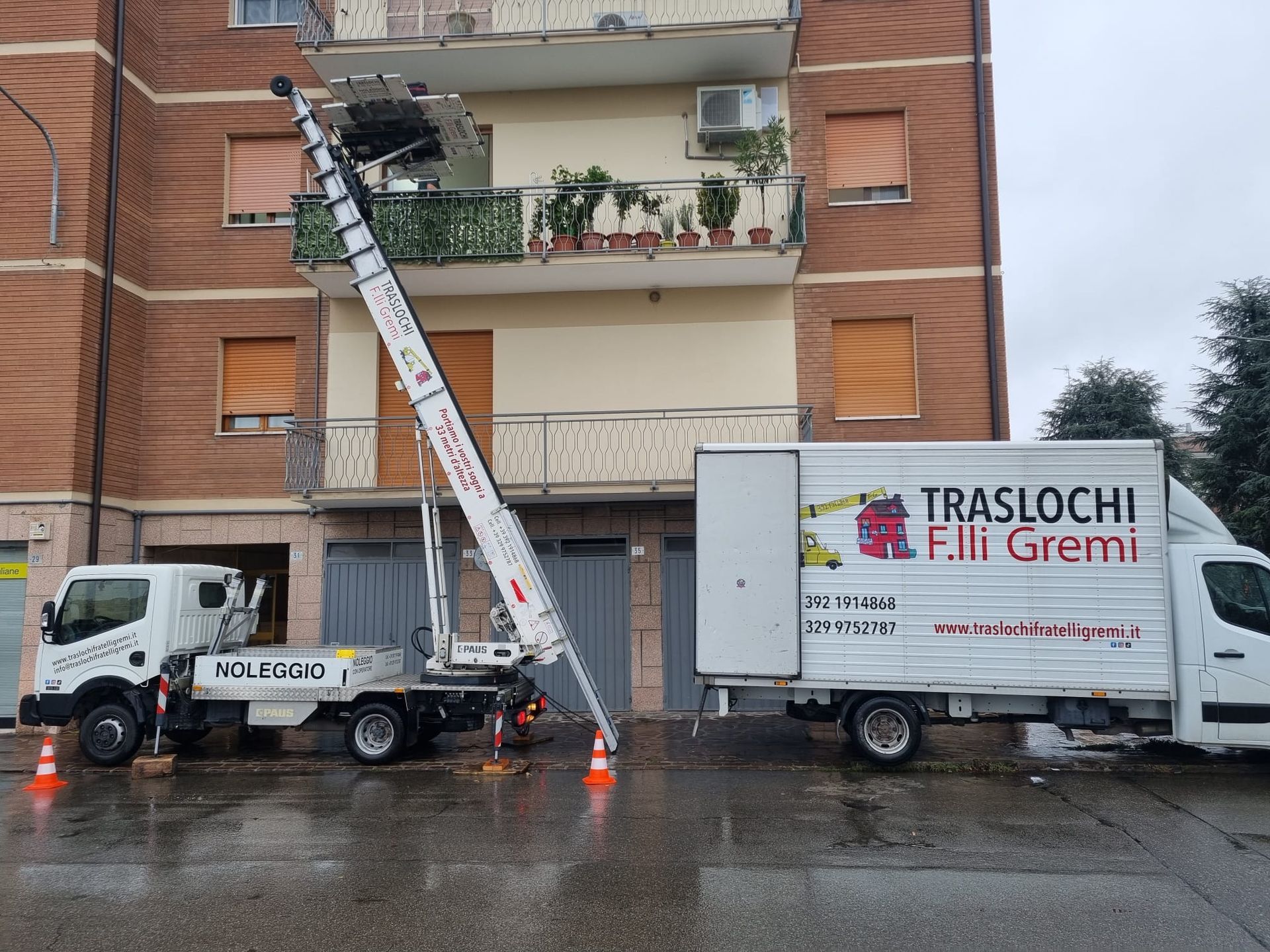 Camion per traslochi con piattaforma elevatrice a scala davanti a un condominio in mattoni su una strada bagnata.