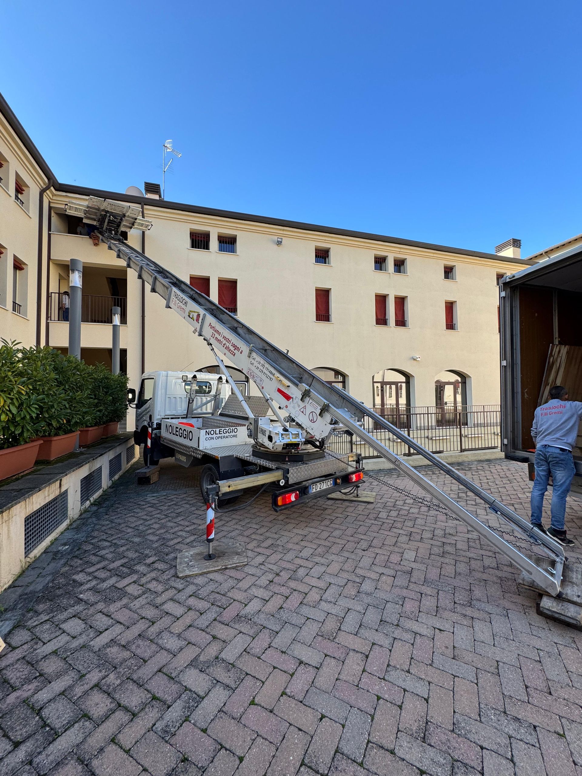 Piattaforma elevatrice da cantiere parcheggiata in un cortile accanto a un edificio beige sotto un cielo azzurro limpido.