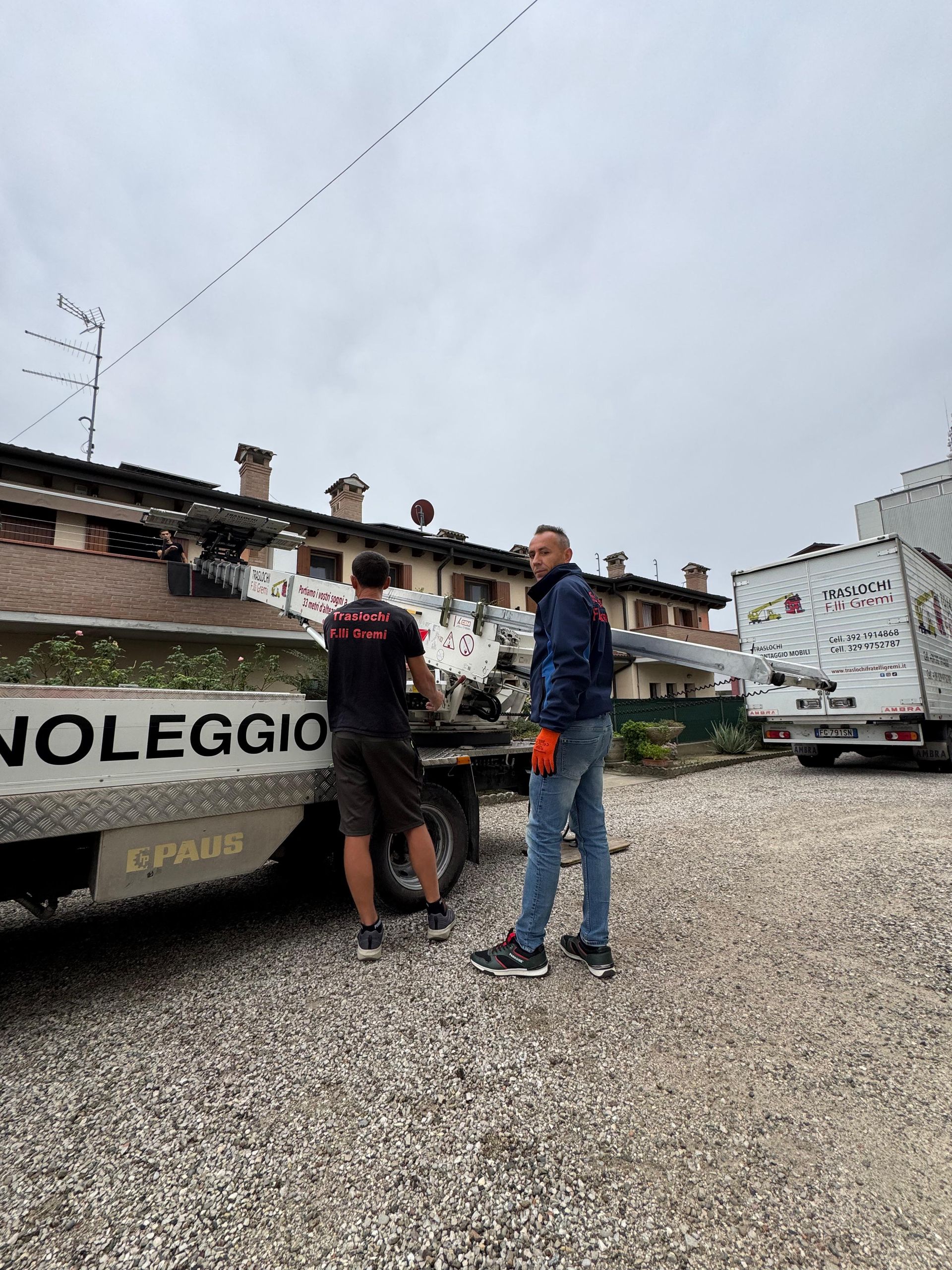 Due persone in piedi accanto a un camion bianco in un piazzale ghiaioso, con edifici e veicoli parcheggiati alle loro spalle.