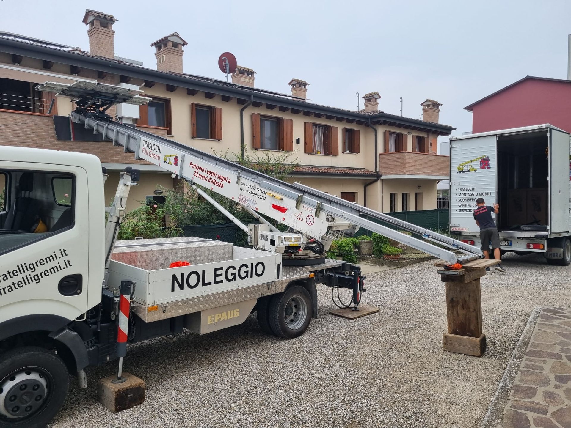 Un carro attrezzi carica un'auto su un pianale in un piazzale ghiaioso, con un edificio e un rimorchio nelle vicinanze.