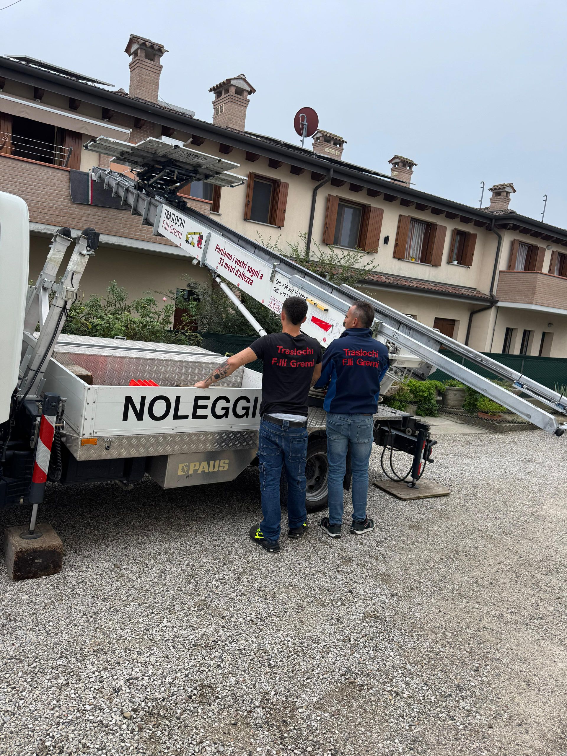 Operai accanto a un camion bianco con una piattaforma aerea montata, in un deposito di ghiaia, vicino a un edificio con persiane in legno.