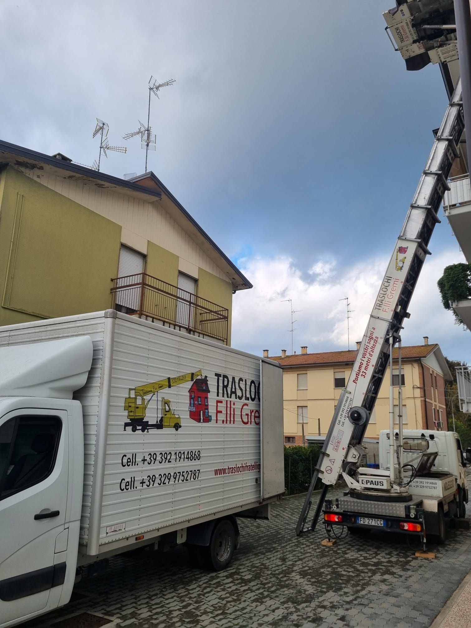 Un camion bianco per traslochi è parcheggiato accanto a una casa, con una gru che solleva attrezzature in una strada residenziale.