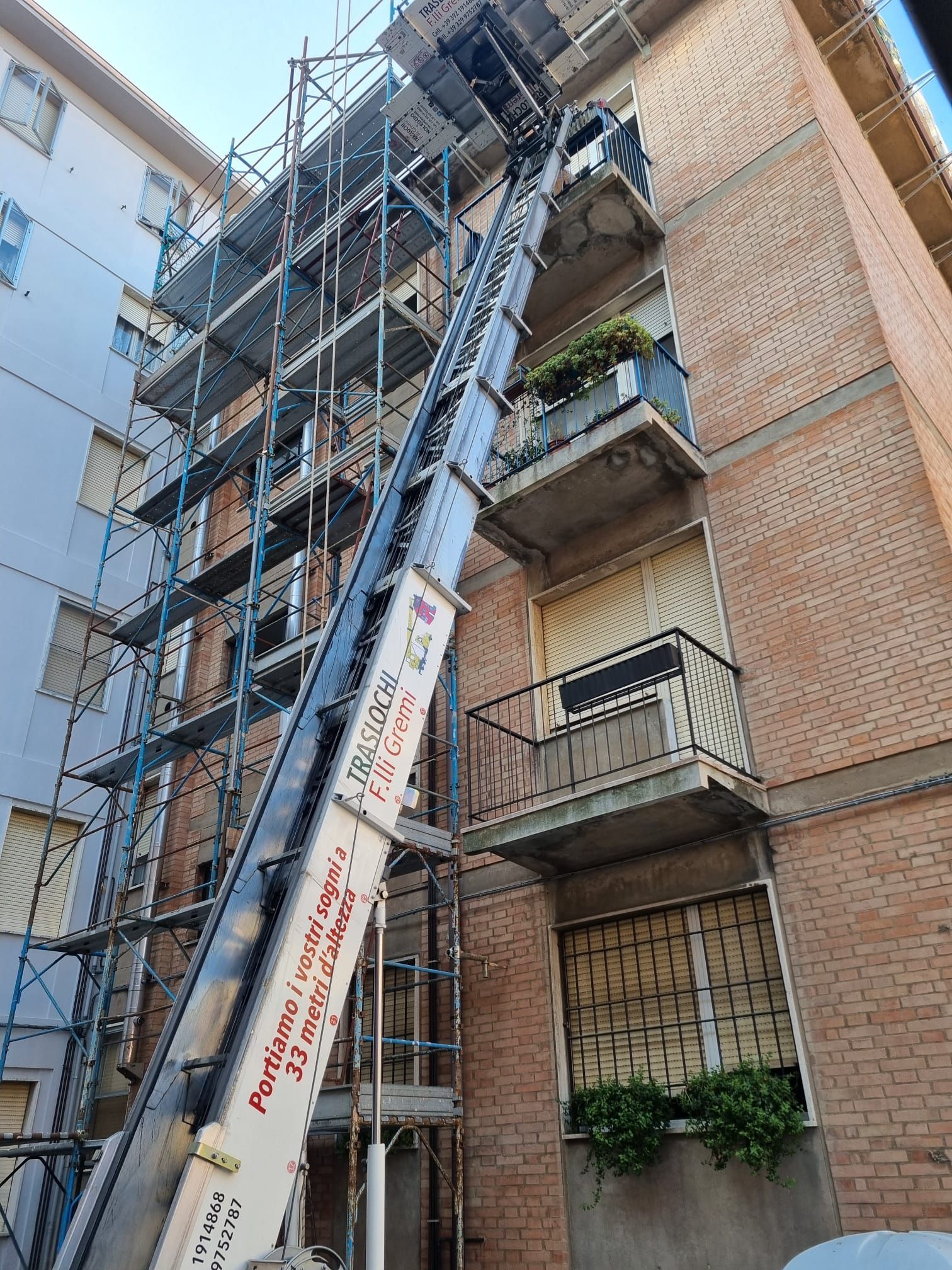 Ponteggio e scala accanto a un condominio in mattoni con balconi e piante in vaso.