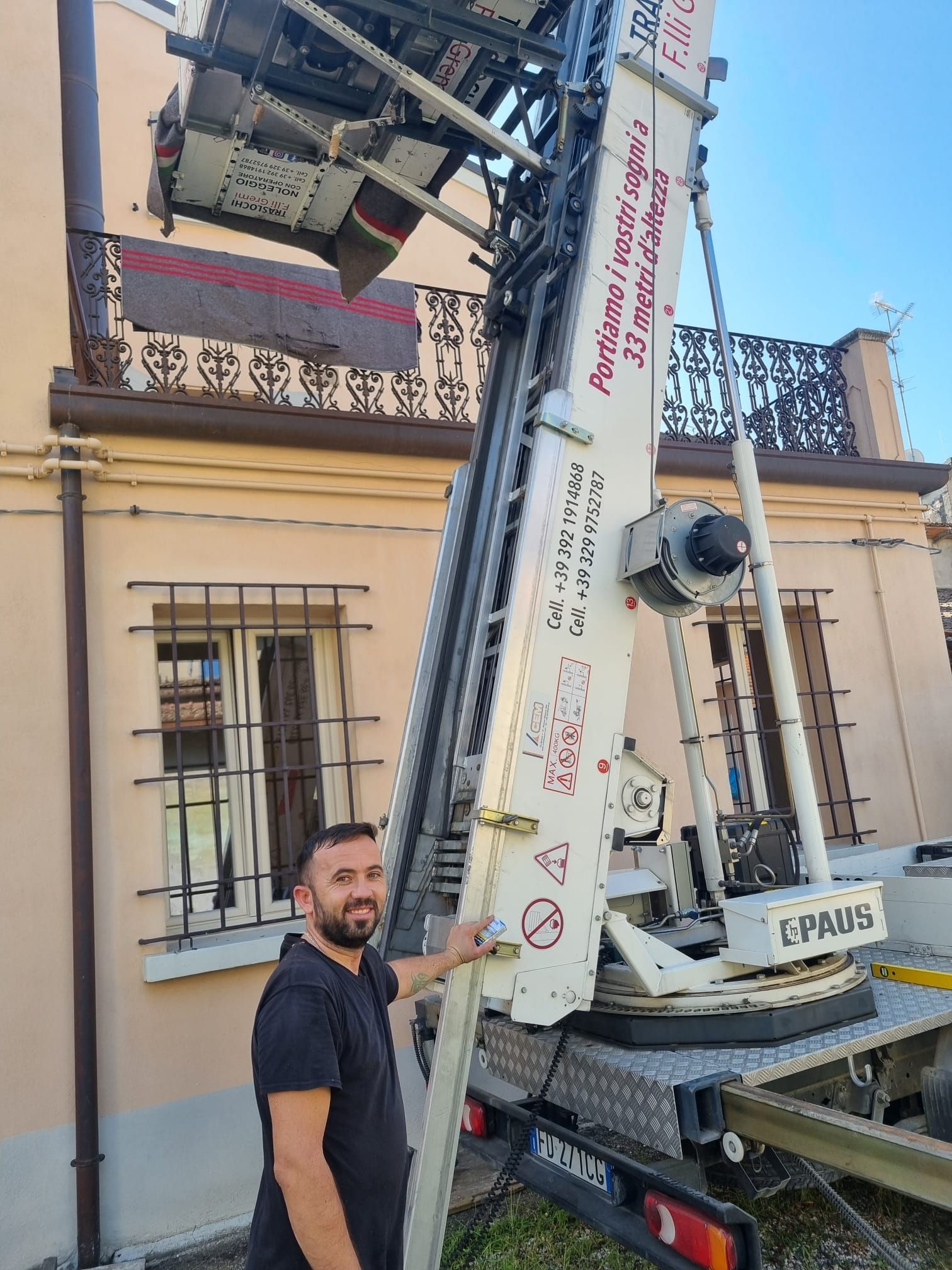 Un uomo è in piedi accanto a una gru bianca che solleva attrezzature vicino a un edificio beige con un balcone.