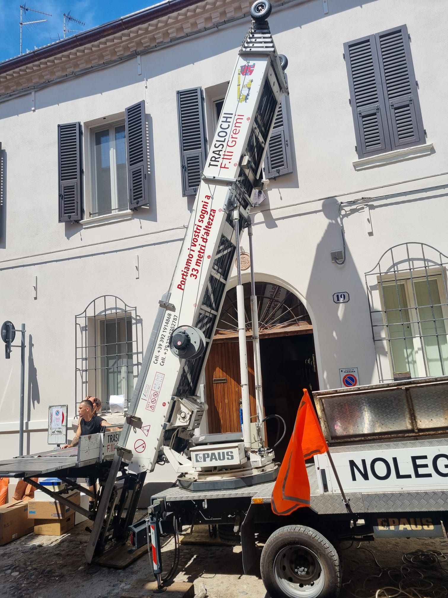 Un camion da lavoro e un'attrezzatura di sollevamento con gru accanto a un edificio bianco con persiane blu.