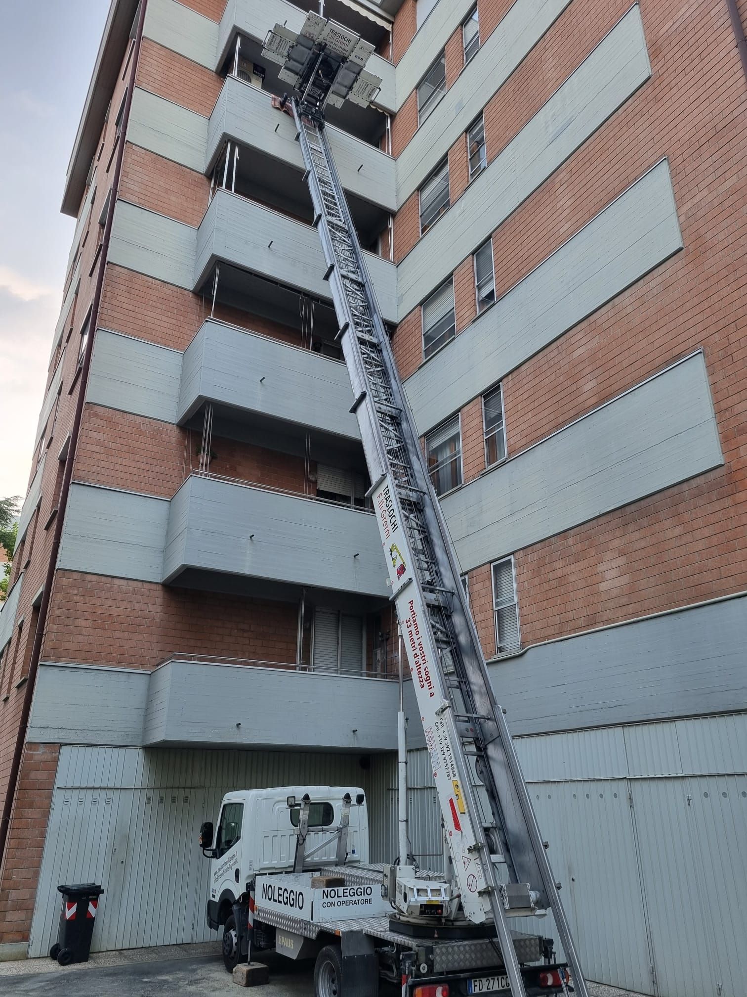 Camion per traslochi con piattaforma elevatrice a scala che raggiunge i piani superiori di un condominio in mattoni.