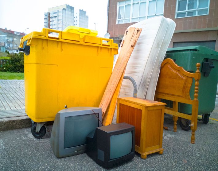 Mobili, materassi e televisori scartati sono ammucchiati accanto a grandi bidoni gialli e verdi all'aperto.
