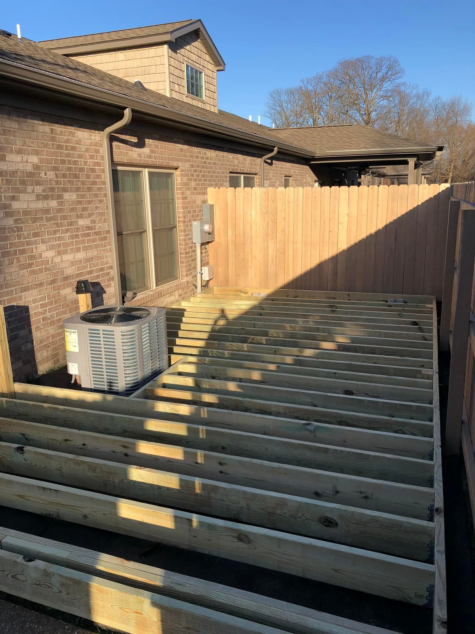 Unfinished wooden deck next to a brick building and a wooden fence; air conditioning unit on the deck.