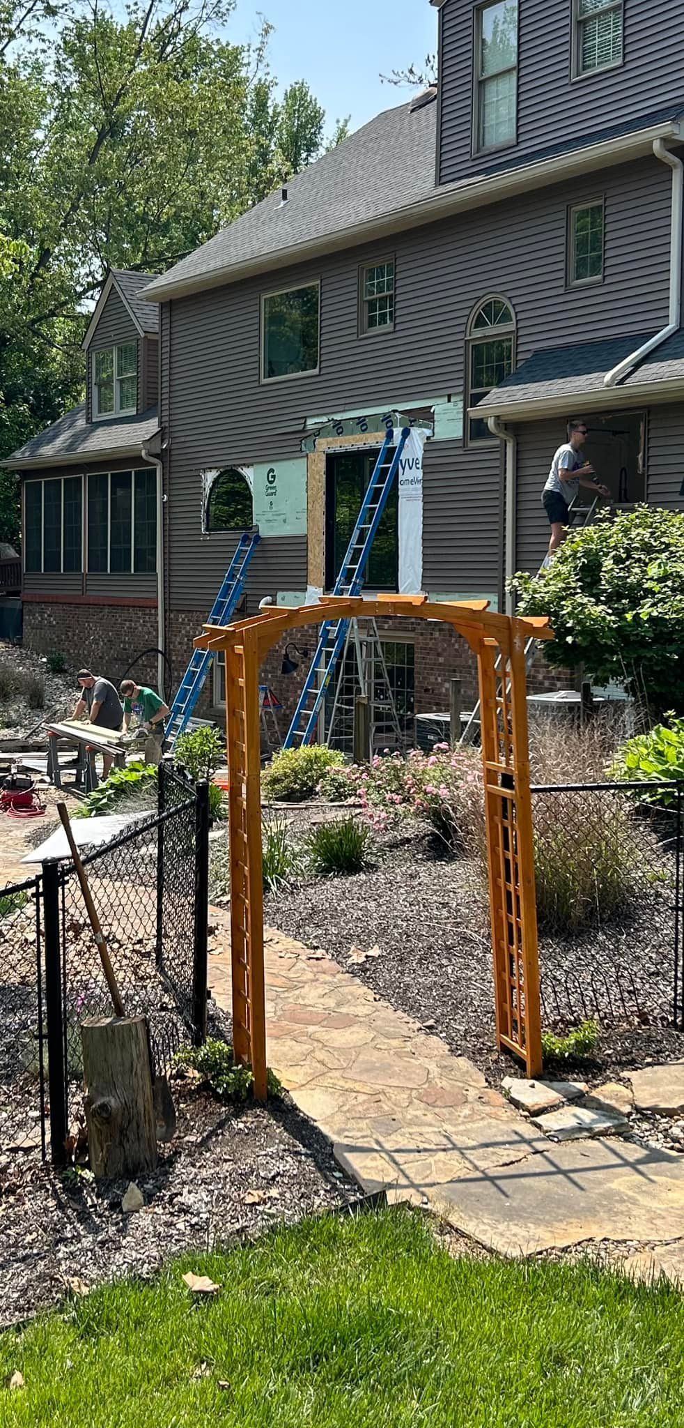 Wooden archway over a stone path leading to a house with a blue ladder and lush greenery.