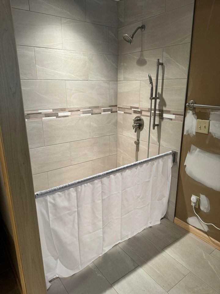 Shower stall with beige tile, shower head, and white curtain.