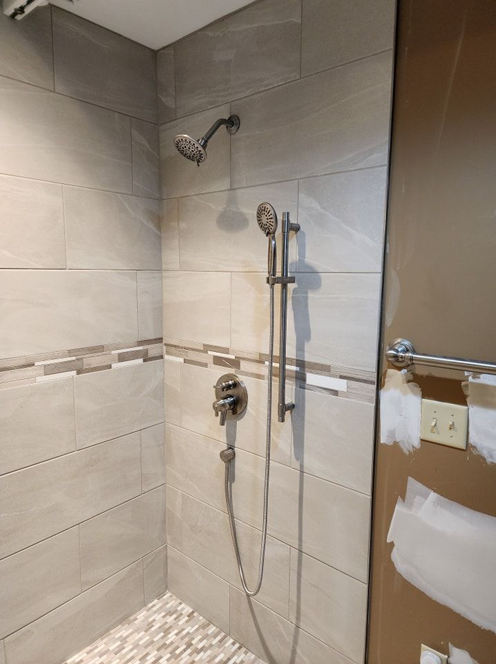 Shower with gray tiled walls, pebble floor, and silver fixtures. Includes a handheld shower head and controls.